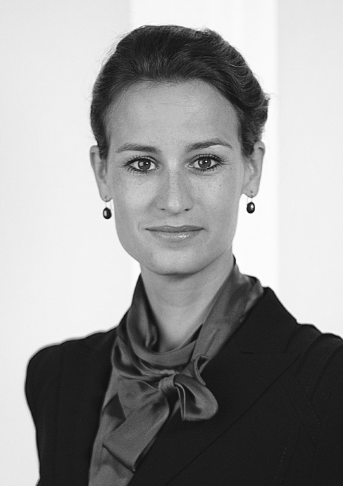 Eine Frau mit dunklem, nach hinten gebundenem Haar, einem dunklen Blazer, einem Seidenschal und Tropfenohrringen blickt in die Kamera und lächelt leicht. Das Schwarz-Weiß-Foto hat einen schlichten Hintergrund, der sie als selbstbewusstes Teammitglied hervorhebt.