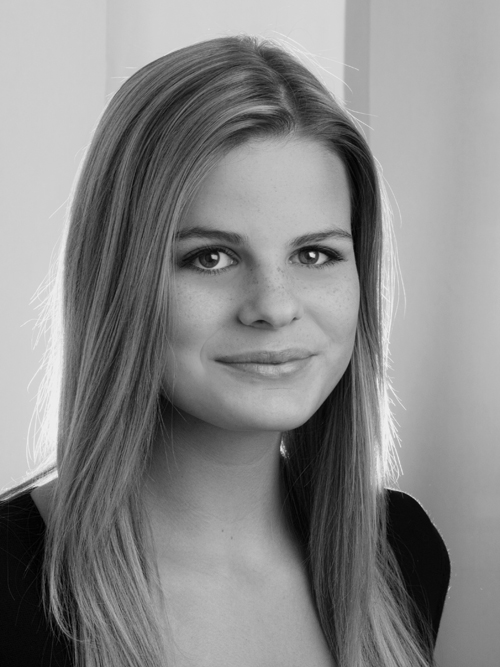 Eine junge Frau mit glattem, langem Haar und heller Haut lächelt sanft. Sie trägt ein dunkles Oberteil und ist in Schwarz-Weiß vor einem sanft beleuchteten Hintergrund fotografiert, der die Wärme einfängt, die sie in das Team bringt.