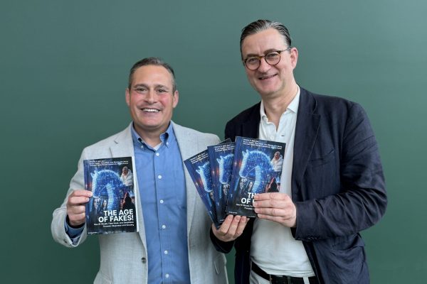 Two men are smiling and holding copies of The Age of Fakes while standing in front of a plain green background.